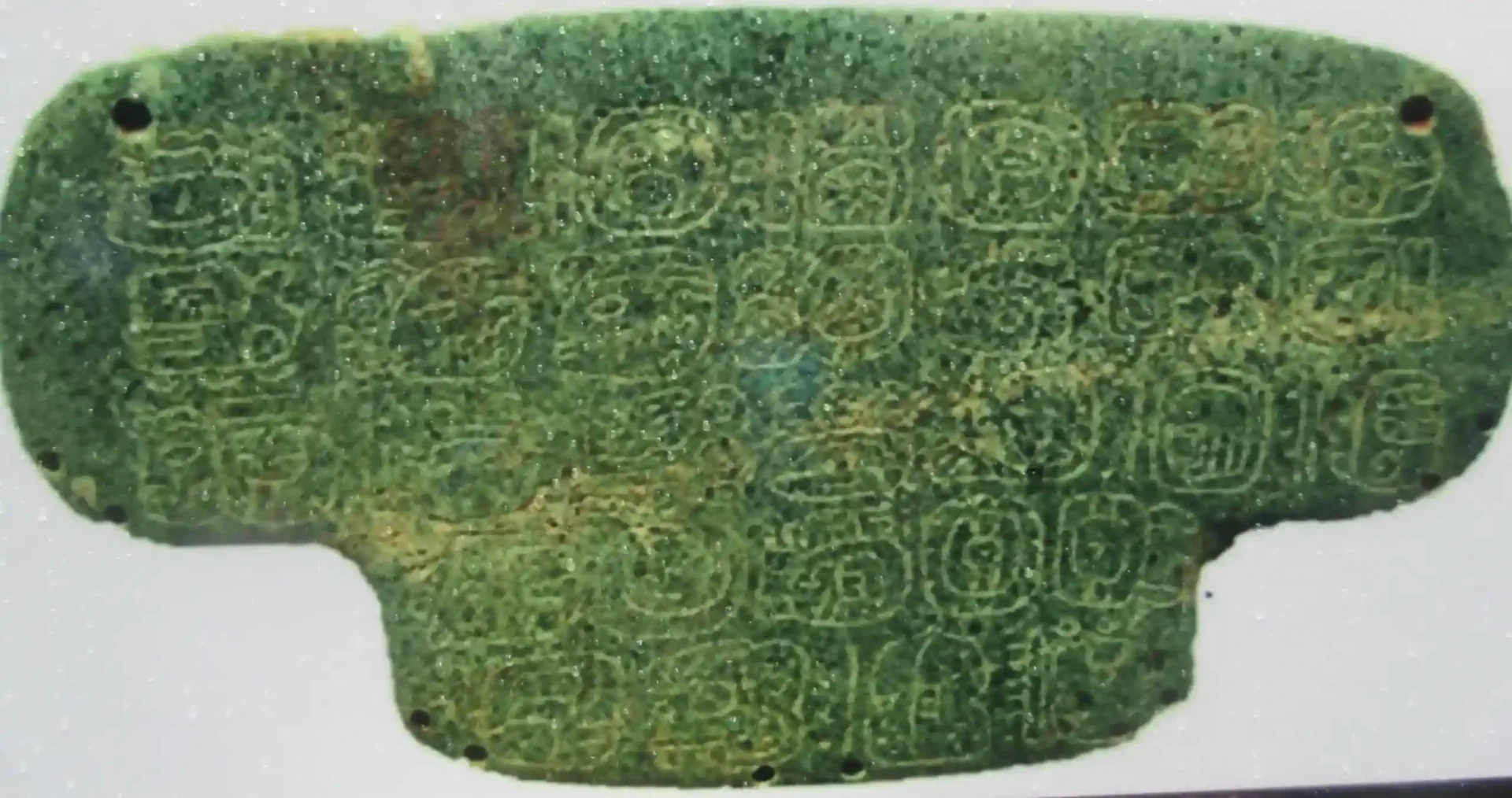The Chocolate & Nim Li Punit Maya Site is an ancient green stone tablet adorned with what might be hieroglyphic symbols in rows. Its worn surface and slightly irregular edges suggest age and historical significance.