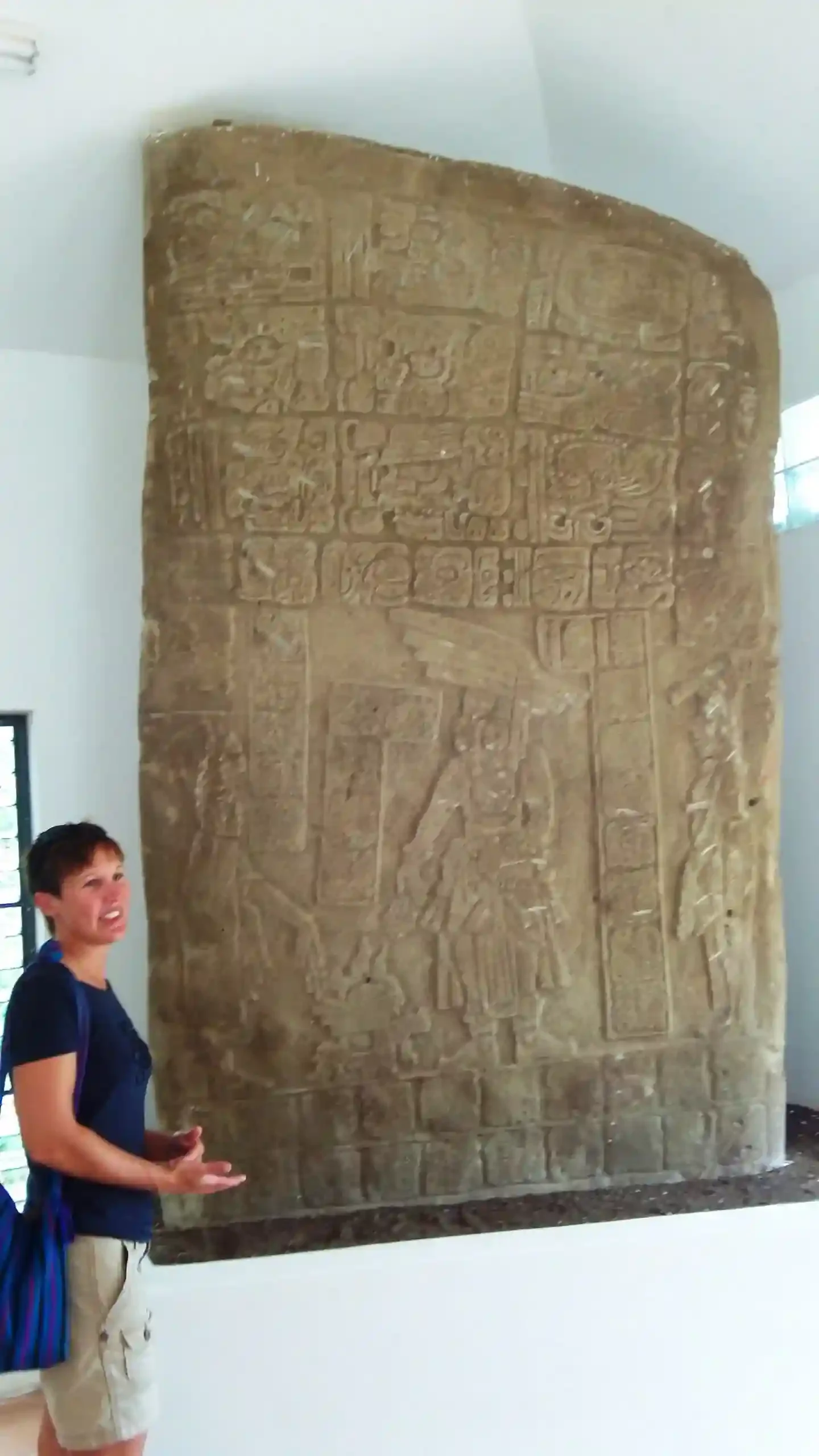In a museum setting, a person stands beside the Chocolate & Nim Li Punit slab, noted for its intricate ancient carvings.