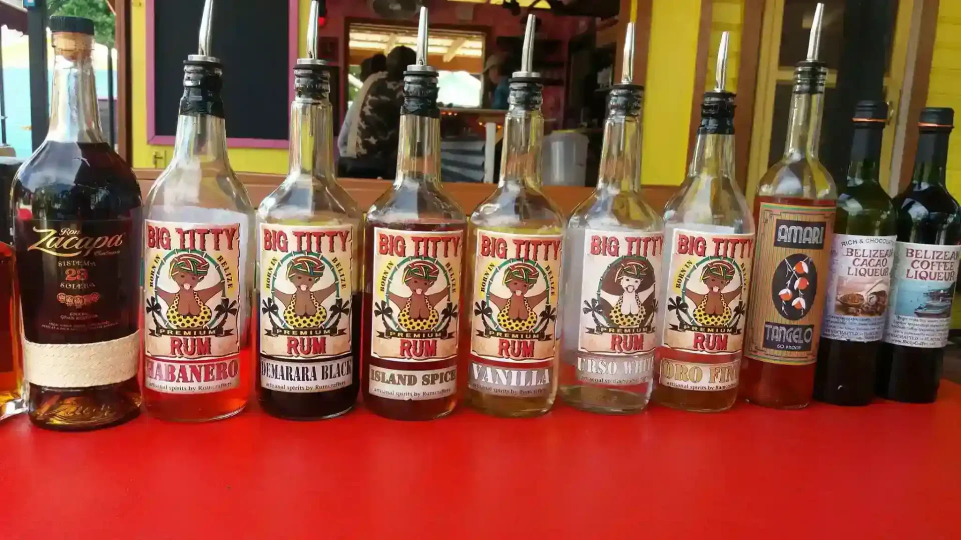 A line-up of various rum bottles on a red surface, each with a differently labeled "Big Titty Rum" design, alongside a few other liquor bottles.