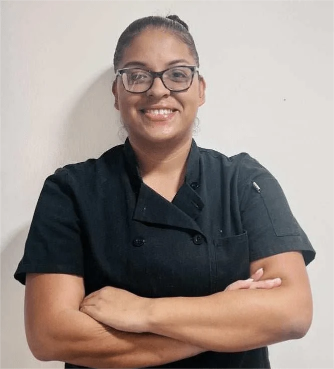 The individual with glasses, dressed in a black chef uniform from "Import placeholder for 2830," stands against a plain background, arms crossed and smiling.
