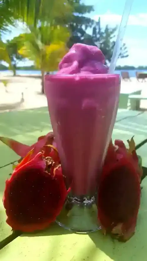 A glass of bright pink smoothie with a straw, surrounded by halved dragon fruits, set on a green table against a beach backdrop with trees and sand.
