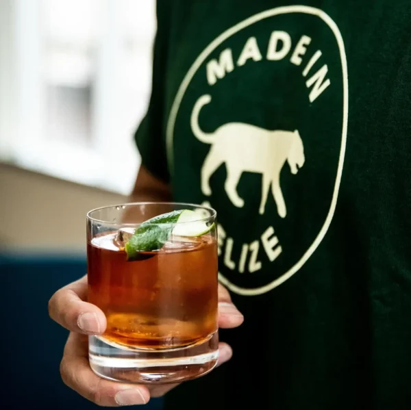 A person wearing a green "Made in Belize" shirt holds a glass of brown cocktail garnished with a lime wedge.