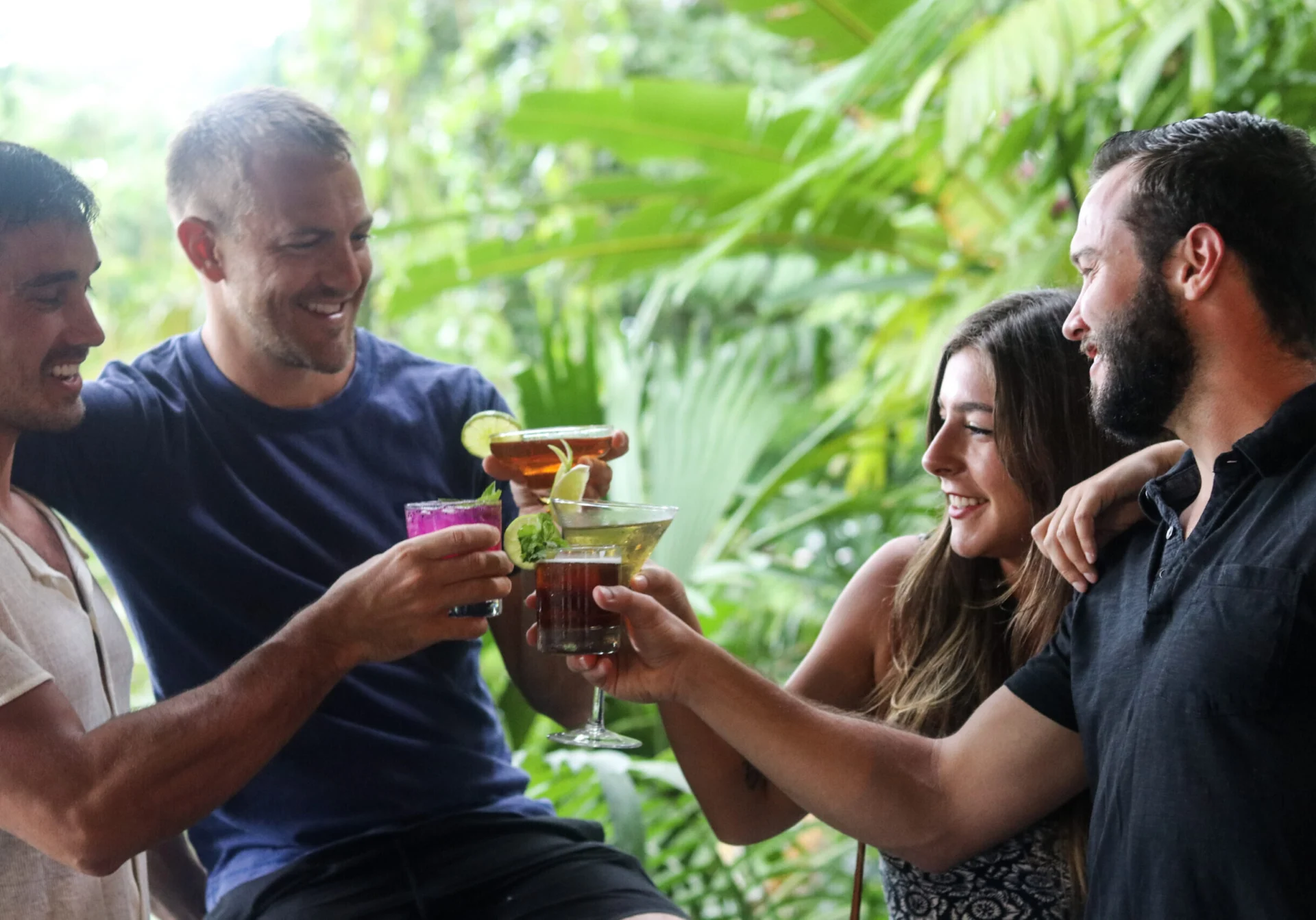 Four people enjoying the "Explore Rum! A Private Rum Class" outdoors clink glasses amidst lush greenery during a casual gathering.