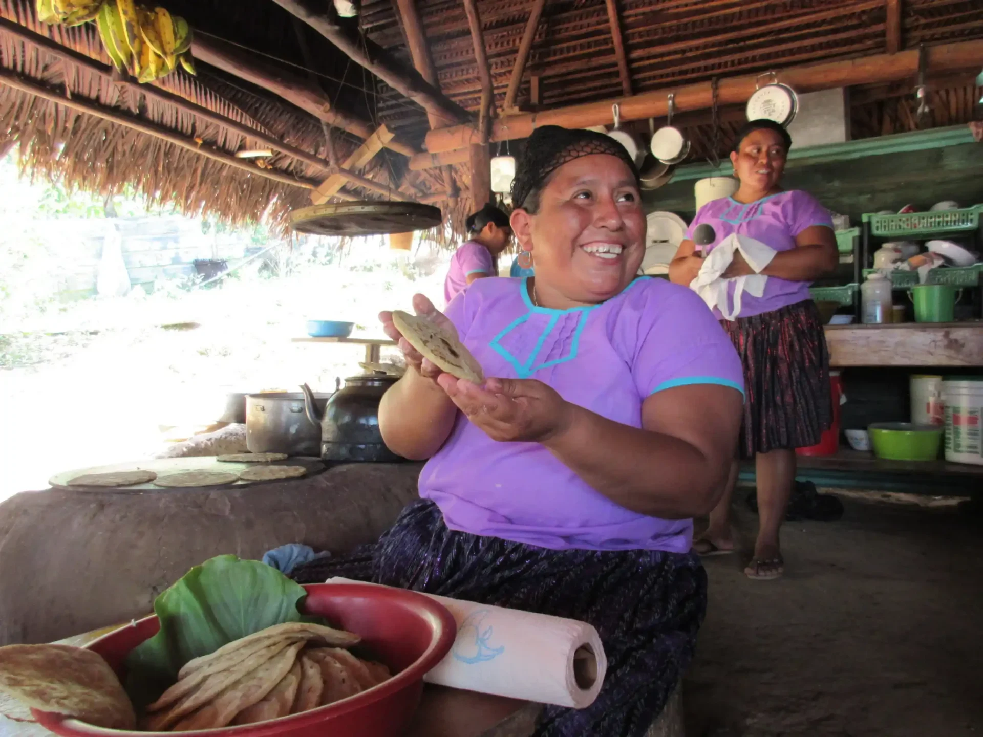 A woman in a purple shirt smiles while making tortillas in a rustic kitchen, with the scene inspired by Maya Fire: Maya Cooking and Adventure. Another woman and various kitchen items appear in the background.