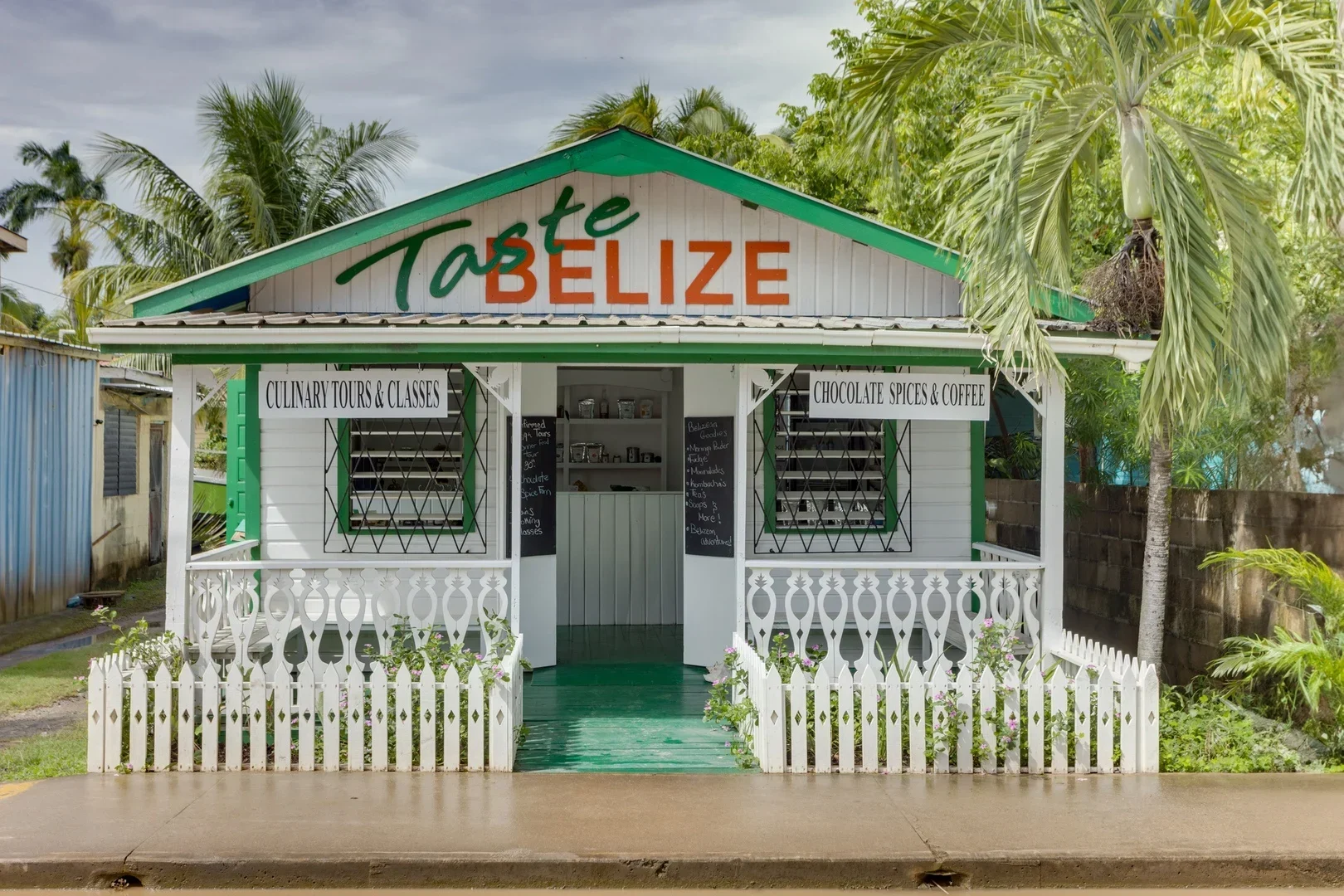 A small, white and green shop labeled "Taste Belize" with signs for culinary tours and chocolate, spices, and coffee, surrounded by palm trees.