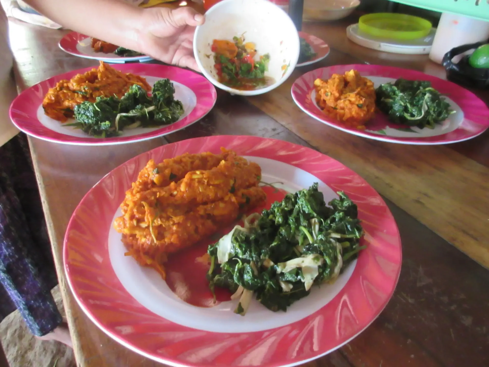 A bowl with a small portion of food is held over a table set with plates, including leafy greens and an orange dish, echoing the flavors of the 3 Adventures Combo Tour.