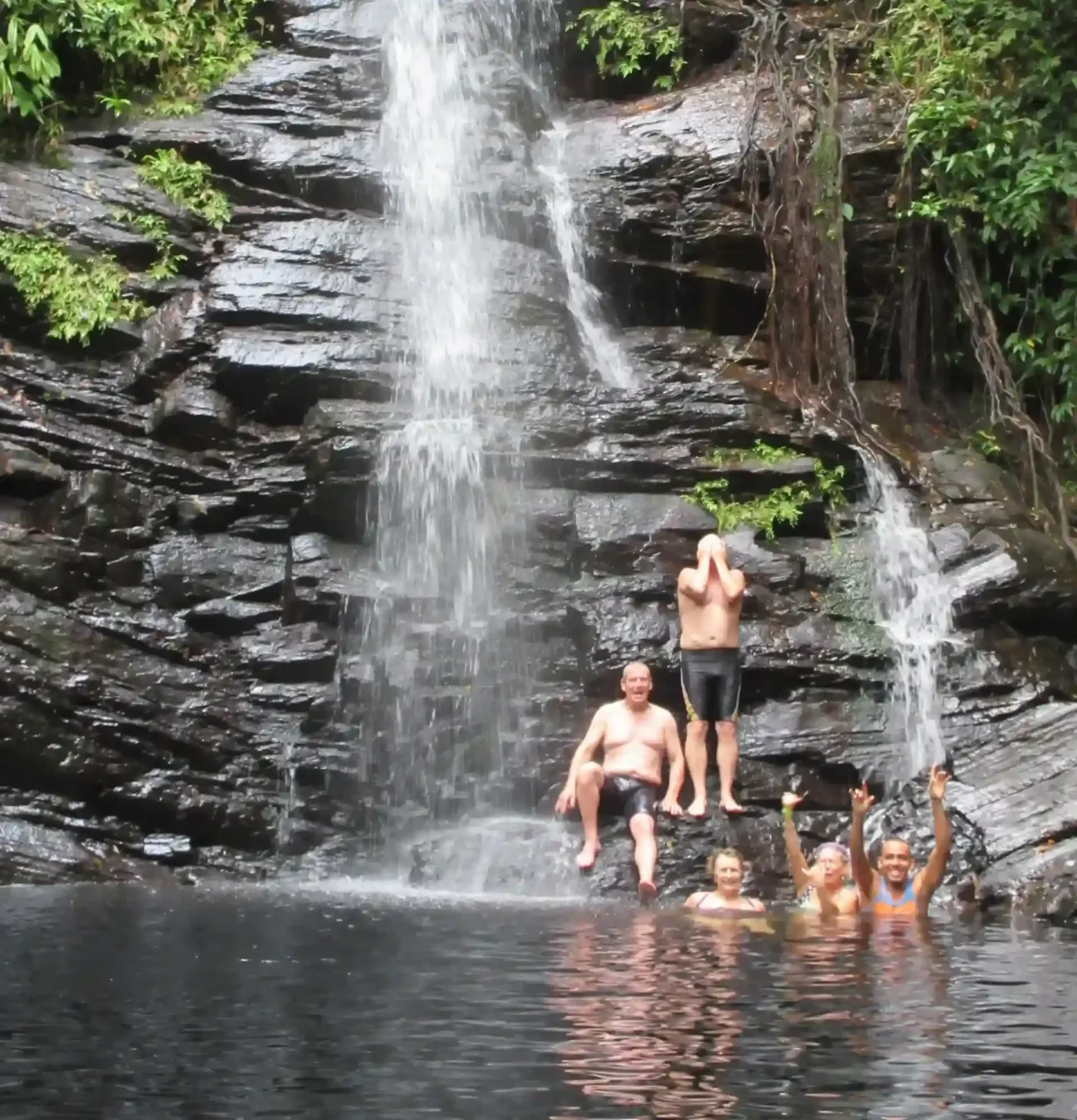 Five people in swimsuits pose and wave in a natural pool at the base of a rocky waterfall surrounded by greenery.