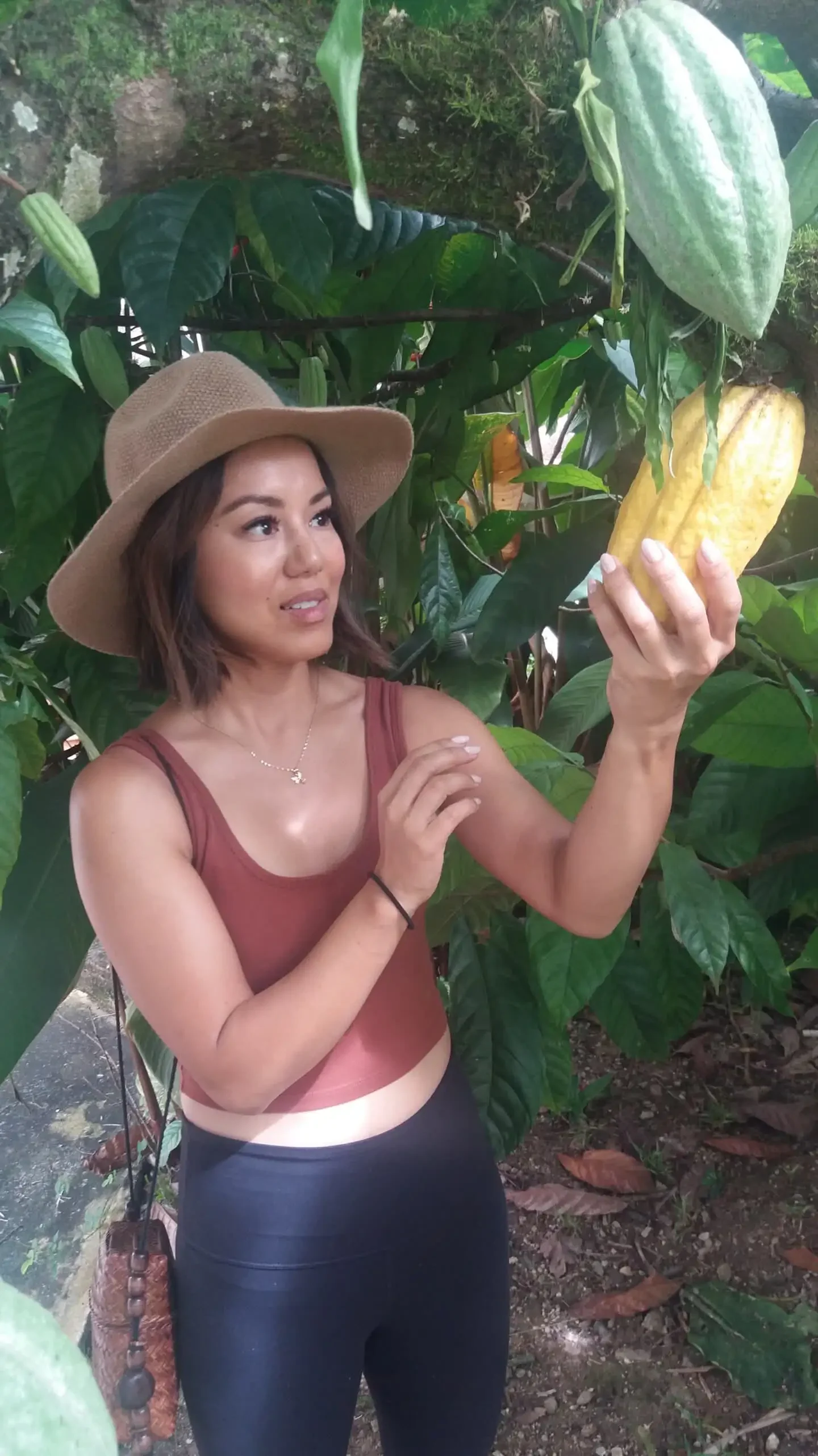 A woman in a hat holds and examines a large yellow cacao pod amidst the lush greenery of the Marie Sharps Hot Sauce Combo Tour garden.