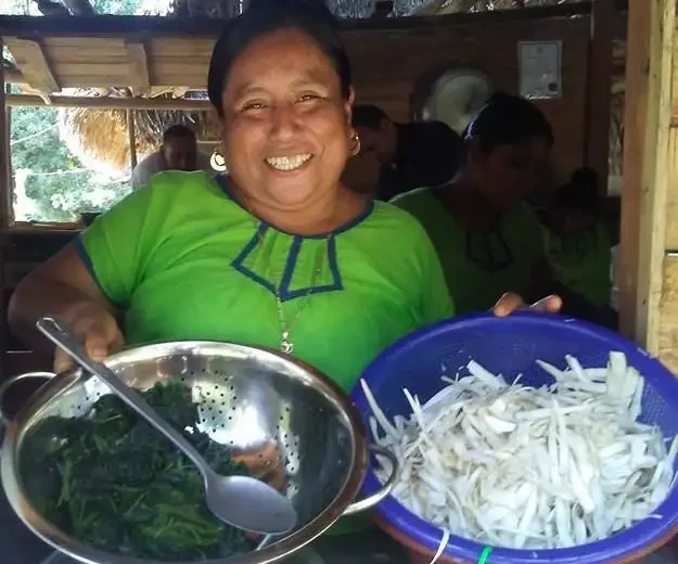 A woman wearing a green blouse holds a silver colander full of greens and a blue basket of sliced white vegetables, smiling indoors as part of her 3 Adventures Combo Tour experience.