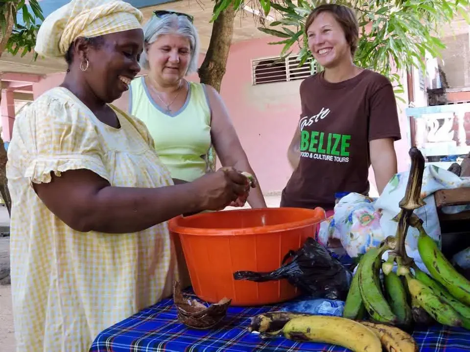 Three women gather around a table outdoors covered with a checkered cloth. One slices plantains over a bowl, while the others watch and smile, with a bottle from the Marie Sharps Hot Sauce Combo Tour set nearby.