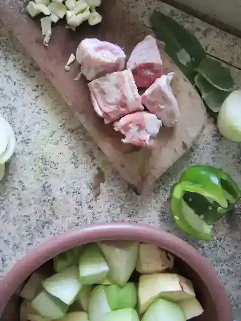 Ingredients on a countertop: raw meat chunks on a cutting board, sliced garlic, onions, bell peppers, bay leaves, and a bowl with chopped apples.