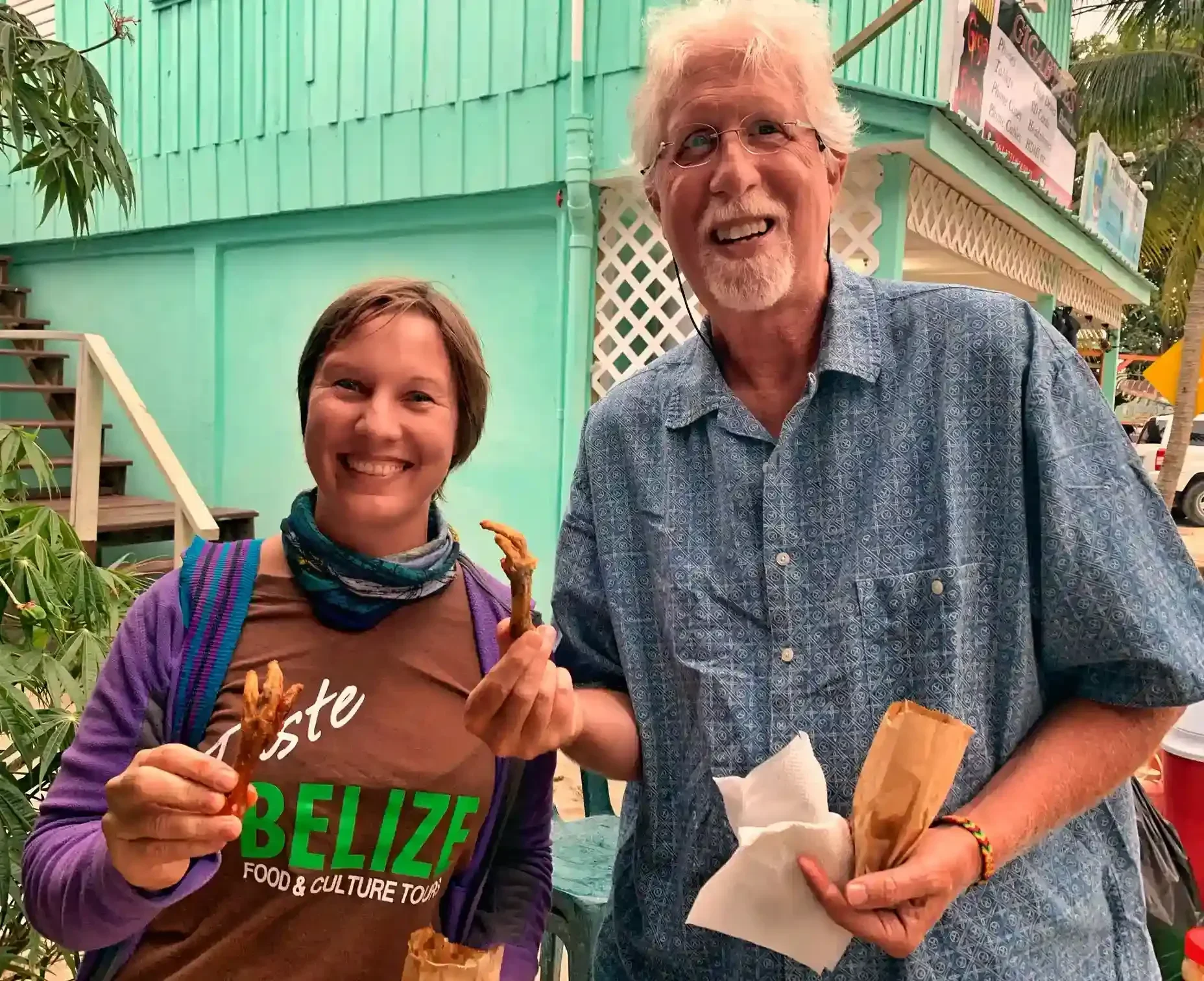 Two people smiling and holding food in their hands, standing outdoors in front of a teal building. One person is wearing a "Taste Belize" shirt.