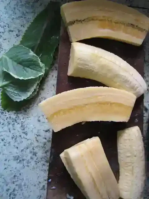 Peeled plantains cut into pieces on a wooden board with two green leaves on a speckled countertop.