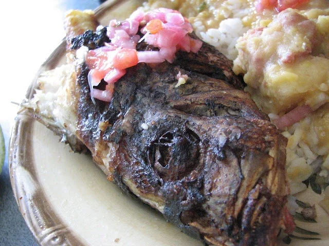 A close-up of a grilled fish head topped with chopped onions and tomatoes, served on a plate with rice and vegetables.